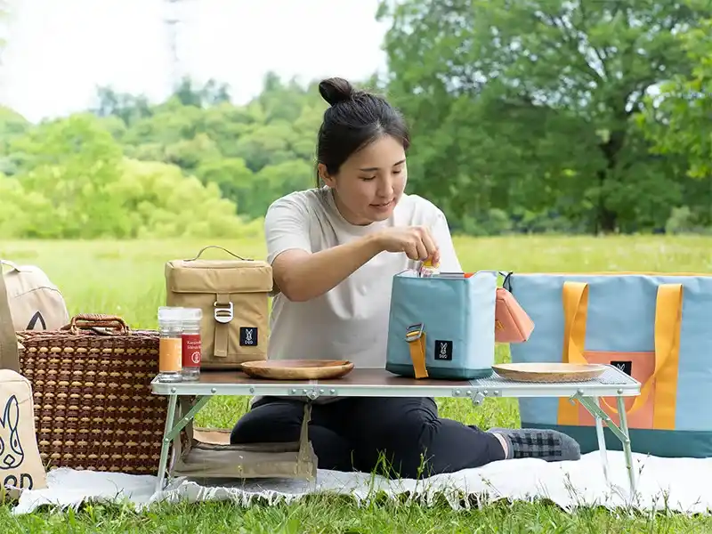 公園でレジャーシートの上に置いたテーブルから、DOD「ヒャドチョミーレ」を使って調味料を取り出す女性の様子。