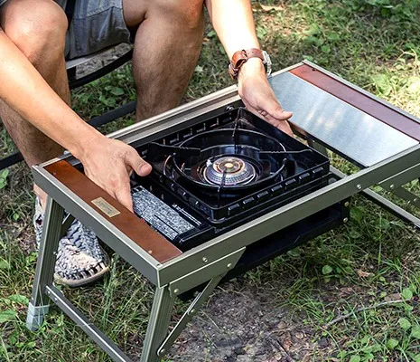 タフまる対応のコンロラック付きIGTテーブルに、カセットコンロ「タフまる」をセットしようとしているキャンプシーン。