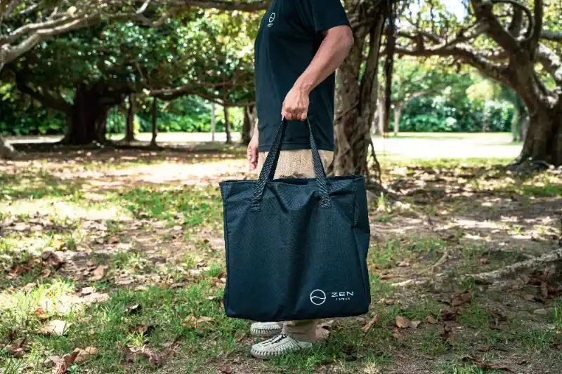 キャンプサイトでZENキャンプ(ZEN Camps)の『FoldFlex Table』を収納バッグに入れて片手で持っているサイズ感が分かる写真